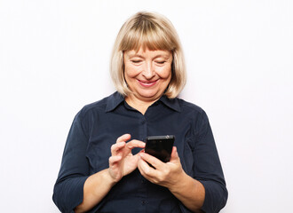 Pretty senior female in casual outfit using modern smartphone and laughing out loud while standing on white background. Old woman watching funny videos on internet.