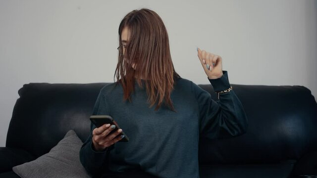 The portrait of a red-haired, freckled girl with glasses looks at the phone screen and performs relaxed dance moves. Dressed in a dark green blouse, sitting on a black sofa. The concept of the woman