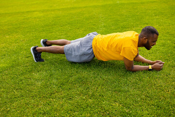 Fototapeta na wymiar Side view of concentrated afro american athlete doing plank exercise on a grass field. Young handsome male working out hard, doing morning training outdoors in yellow sportswear during epidemic 