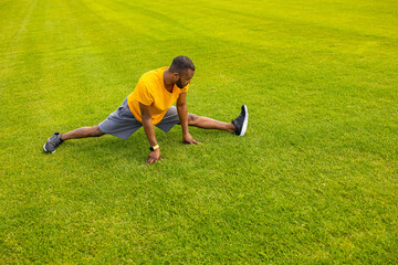 Fototapeta na wymiar Concentrated athletic man in stylish sportswear trying to do a split, sitting at the stadium stretching legs. Sportive african american male working out, doing warm up before training outdoors concept