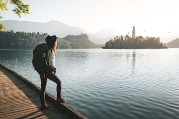Hipester young woman with backpack enjoying breathtaking  sunrise by the lake. Woman wearing hat...