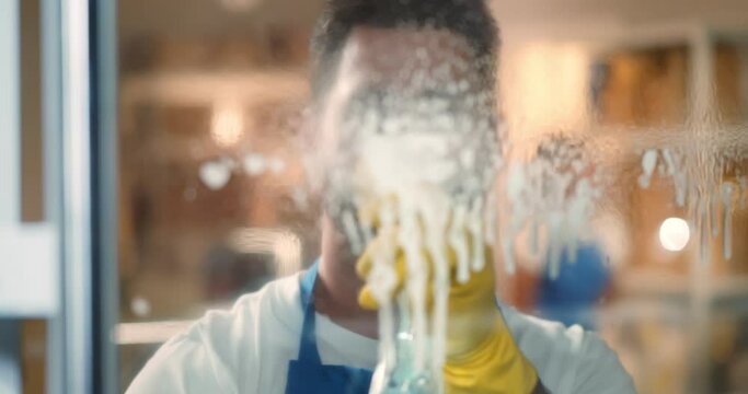 Male janitor using spray and rag to clean window in office wearing apron
