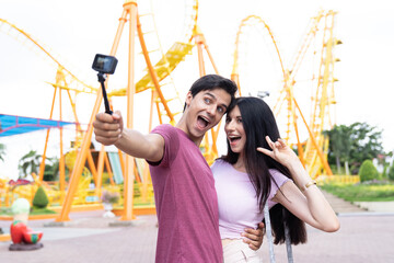 Obraz premium Cheerful caucasian young couple having fun while talking selfie at the amusement theme park on Roller coaster background. Attractive happy couple at the theme park. couple, people, holiday concept