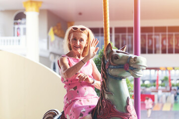 Happy cheerful smiling elderly woman at the amusement park. Senior woman on horse carousel ride at...