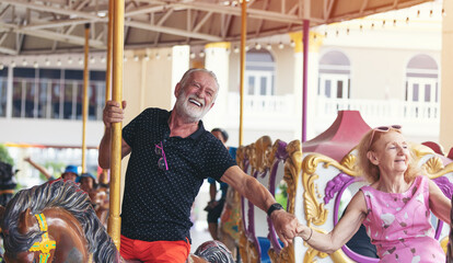 Happy cheerful smiling senior couple holding hands together and playing on horse carousel ride at...