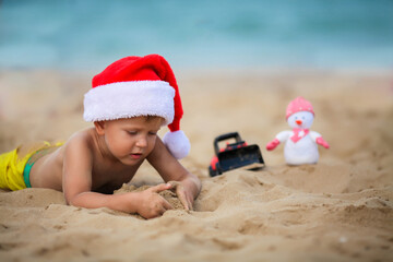 Happy boy in a santa claus hat plays with a toy car on the beach. A child in Christmas is resting on the sea. Copy space