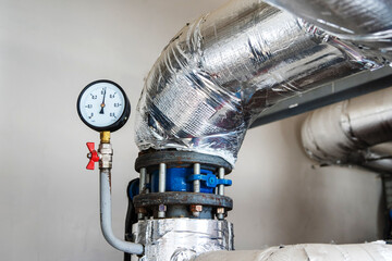 A large-diameter pipe insulated with mineral wool and foil is located in the boiler room of an apartment building. Manometer for monitoring the pressure in the pipeline. Close-up - obrazy, fototapety, plakaty
