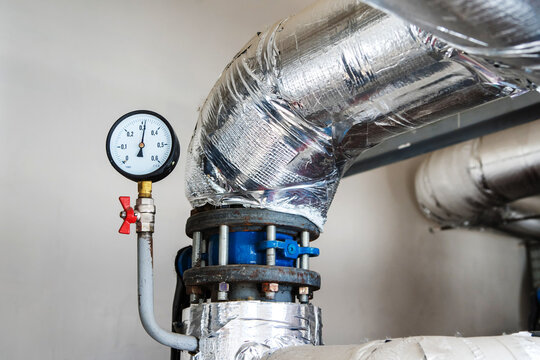A large-diameter pipe insulated with mineral wool and foil is located in the boiler room of an apartment building. Manometer for monitoring the pressure in the pipeline. Close-up