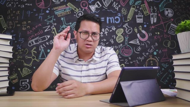 Happy asian teacher glasses man teaching and looking at camera while sitting at desk in classroom. Male teacher explain to student in class via video conference. E-learning, learn from home, education