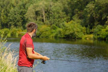 A man with a beard is fishing on the river. A fisherman with a fishing rod is fishing on the river bank.