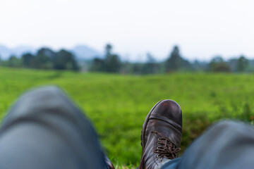 Relaxing on green field with leather boots. Man's leg with brown leather shoes and blue jeans sitting calmly enjoying outdoor landscape view of tea plantation. POV point of view of relax sitting.