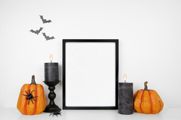 Halloween mock up. Black frame on a white shelf with pumpkin decor and black candles. Portrait frame against a white wall with bats. Copy space.