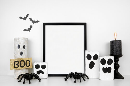 Halloween mock up. Black frame on a white shelf with rustic wood ghost decor, spiders and black candle. Portrait frame against a white wall with bats. Copy space.