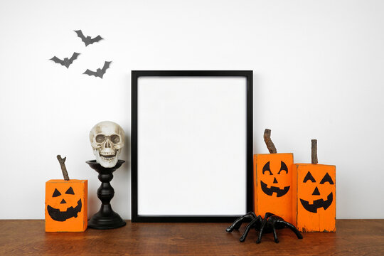 Halloween mock up. Black frame on a wood shelf with rustic wood jack o lantern decor, spiders and skull. Portrait frame against a white wall with bats. Copy space.