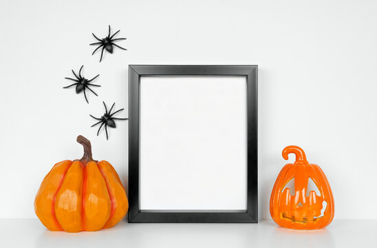 Halloween mock up. Black frame with pumpkin and jack o lantern decor on a white shelf. Portrait frame against a white wall with spiders. Copy space.