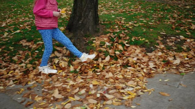 Unknown little girl in white sneakers walks through dry yellow leaves along the path of autumn park, slow motion.