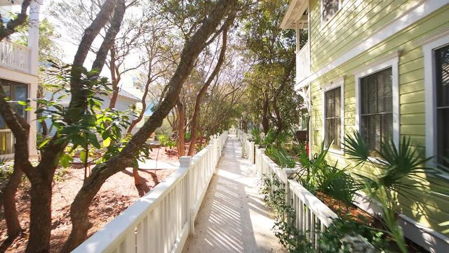 Point of view pov handheld shot walking on small sand street sidewalk between buildings in town of Seaside, Florida with pastel colorful houses new urbanism and garden trees in winter sunny sunlight
