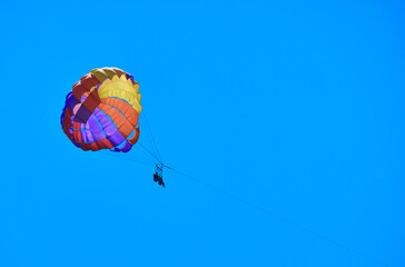 Man flies on rainbow-colored parachute against blue sky.