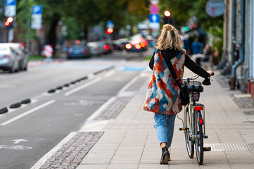woman in brightly colored clothes pushing a bicycle on a sidewalk in the city, view from back