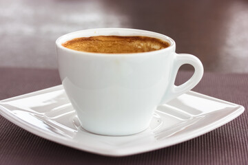 Round white ceramic cup with cappuccino latte coffee brown foam on square saucer on a table. A hot flavored aromatic drink in a cafe, restaurant in the morning, lunch at autumn season. Beverage theme.