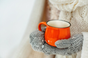 Hands in mittens hold an orange cup. Snowy background in winter 