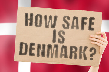 The question " How safe is Denmark ? " on a banner in men's hand with blurred Danish flag on the background. Safety. Street. Outdoor. Dangerous. Security. Attack. Criminal. Criminality