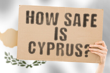 The question " How safe is Cyprus ? " on a banner in men's hand with blurred Cypriot flag on the background. Safety. Street. Outdoor. Dangerous. Security. Attack. Criminal. Criminality