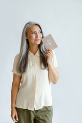 Naklejka premium Thoughtful hoary haired Asian lady customer holds gift card standing on light grey background in studio. Shopping certificate