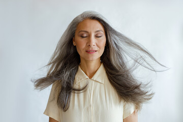 Pretty tranquil Asian woman with flying silver hair stands on light grey background in studio....