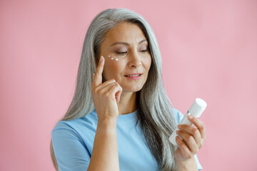 Pretty middle aged Asian woman applies cream under eyes holding bottle on pink background in...