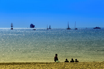 Suggestive sea view from a beach in the city of Genoa with many boats, cargo ships and sailing ships off the coast, against a blue sky. On the sunny beach the silhuettes of some people.. Genoa, Italy.