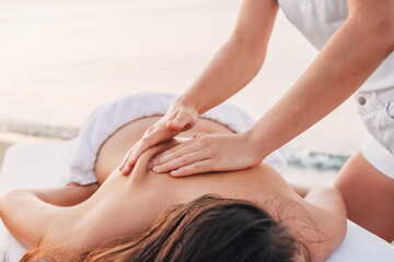 A massage spa girl in white clothes on the seashore makes a back massage to a woman who lies on a massage table.