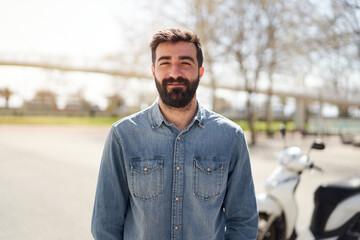 Portrait of a young Spanish man looking at the camera. The man with cheerful smile is in front of his motorcycle, which is parked in the city.