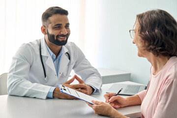 Happy Indian male doctor consulting senior old patient filling form at consultation. Friendly...