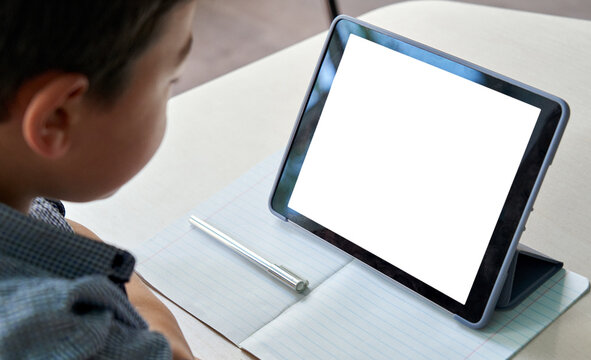 Asian junior school kid girl student using digital tablet looking at white blank empty mockup screen at desk in classroom. Online education, virtual classes software tech ads. Over shoulder view
