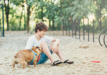 Funny french puppy bulldog and teenager playing games outside. Adorable orange bulldog in blue harness in the playground on a sand. Still life, friendship with a dog. 