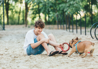 Funny french puppy bulldog and teenager playing games outside. Adorable orange bulldog in blue harness in the playground on a sand. Still life, friendship with a dog. 