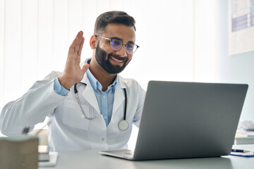 Happy Indian male doctor waving hand having virtual video call online consultation on laptop computer in hospital. Tele health medicine. Telemedicine patients appointment distance remote chat in India - obrazy, fototapety, plakaty