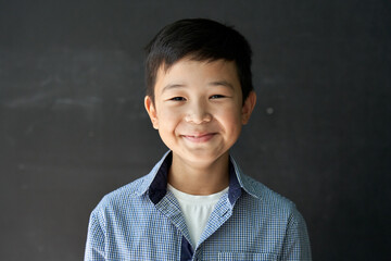 Happy cute Asian kid boy school student looking at camera at blackboard background. Smiling ethnic...