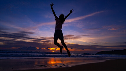 Silhouette of happy kid girl jumping on the beach at sunset or sunrise sky over sea.