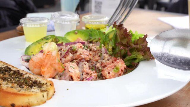 Fresh seafood ceviche mixed with fork and lettuce on side. Chopped salmon, prawns and avocado slices on white plate. Gourmet restaurant food concept