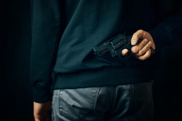 Criminal person with revolver on his back. Man in dark clothing is holding gun. Firearm for defense or attack. Murderer in black background.