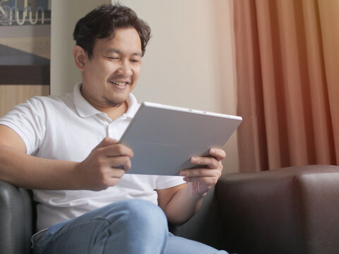 Happy confident young Asian male freelancer reading news on his tablet while sitting on couch in living room, young digital entrepreneur enjoys working from home, modern cozy life 