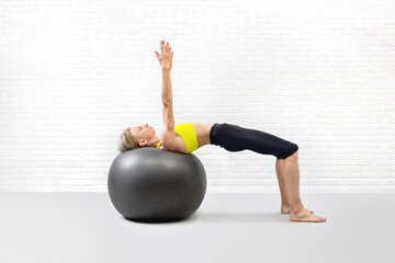 Slim athletic caucasian woman practice shoulder bridge drill using big exercise ball in fitness studio indoor, arms up.