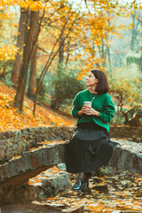 young pretty woman sitting on little stone bridge at city public park enjoying autumn weather drinking coffee