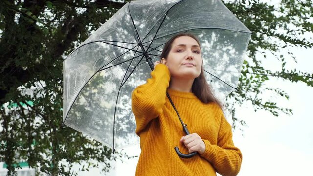 sad woman in a mustard sweater stands and freezes under a transparent umbrella in rainy weather.