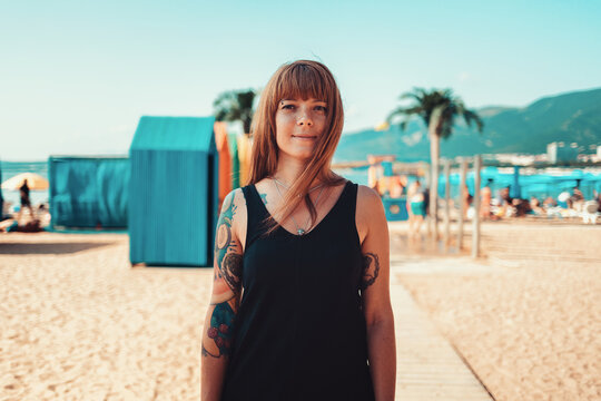 Portrait of a young smiling woman with a tattoo on her arm, looking at the camera. Sandy beach on the background. The concept of summer vacation