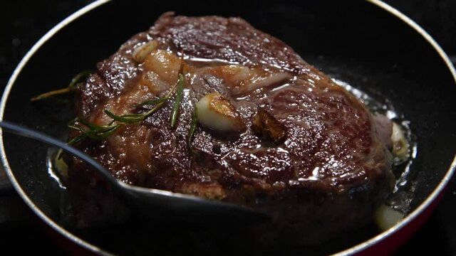 Close-up view of chuck steak frying on frying pan with garlic and rosemary branches. Person pours oil on steak by spoon. Beef meat cooking. Selective focus. 4K resolution video. Food preparation theme