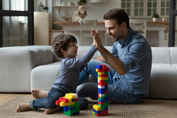 Happy millennial 30s dad giving praise and high five to little son for building toy tower from construction plastic blocks. Joyful kid playing learning games with daddy on heating floor - obrazy, fototapety, plakaty