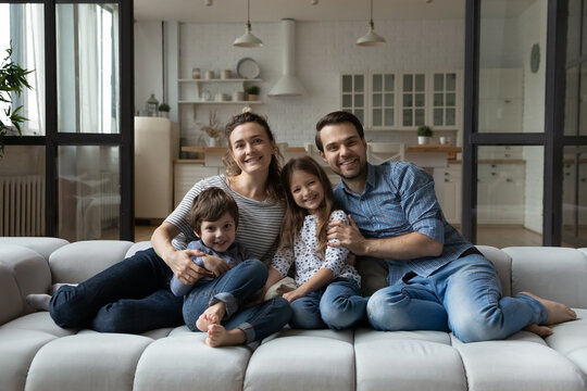 Happy parents and two joyful kids sitting on couch at cozy home, hugging, looking at camera, smiling. Portrait of millennial family couple and children in new apartment. Mortgage, real estate concept